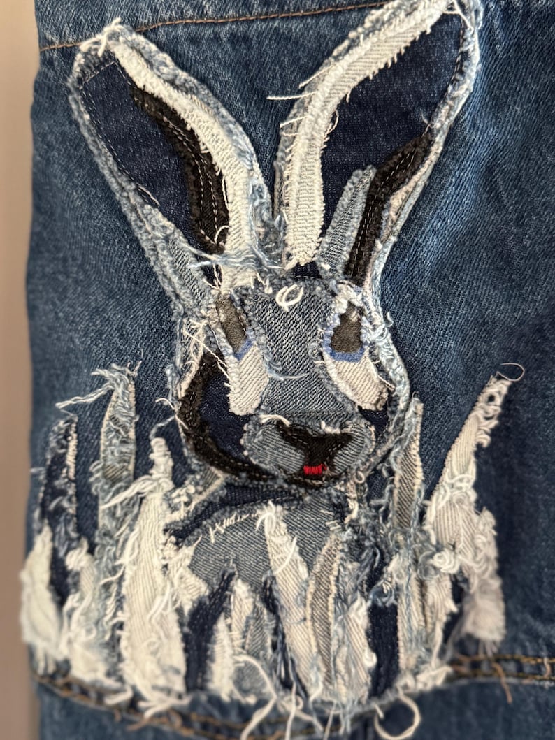 May include: Close-up of a denim jacket detail featuring a rabbit applique. The rabbit is constructed from various denim scraps in shades of blue, white, and gray, with frayed edges. The rabbit has black eyes and a red nose.