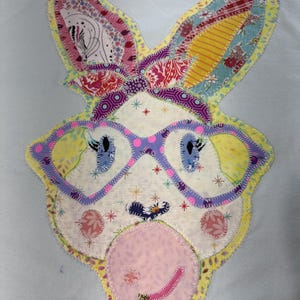 May include: A light blue t-shirt with a patchwork bunny applique. The bunny has pink glasses, a pink nose, and a yellow, pink, and blue patterned headband.
