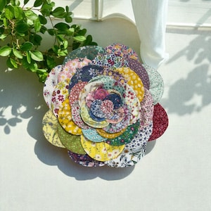 Może przedstawiać: Ręcznie robiony, wielokolorowy kwiat z tkaniny o warstwowym wzorze płatków. Kwiat ma różne wzory kwiatowe w odcieniach żółci, różu, niebieskiego i zieleni. Kwiat prawdopodobnie służy do celów dekoracyjnych.