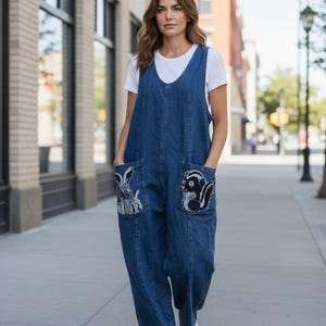 May include: A woman wearing a blue denim jumpsuit with two large front pockets. Each pocket features an embroidered animal design: a rabbit on the left and a skunk on the right. The jumpsuit is paired with a white t-shirt and white sneakers.