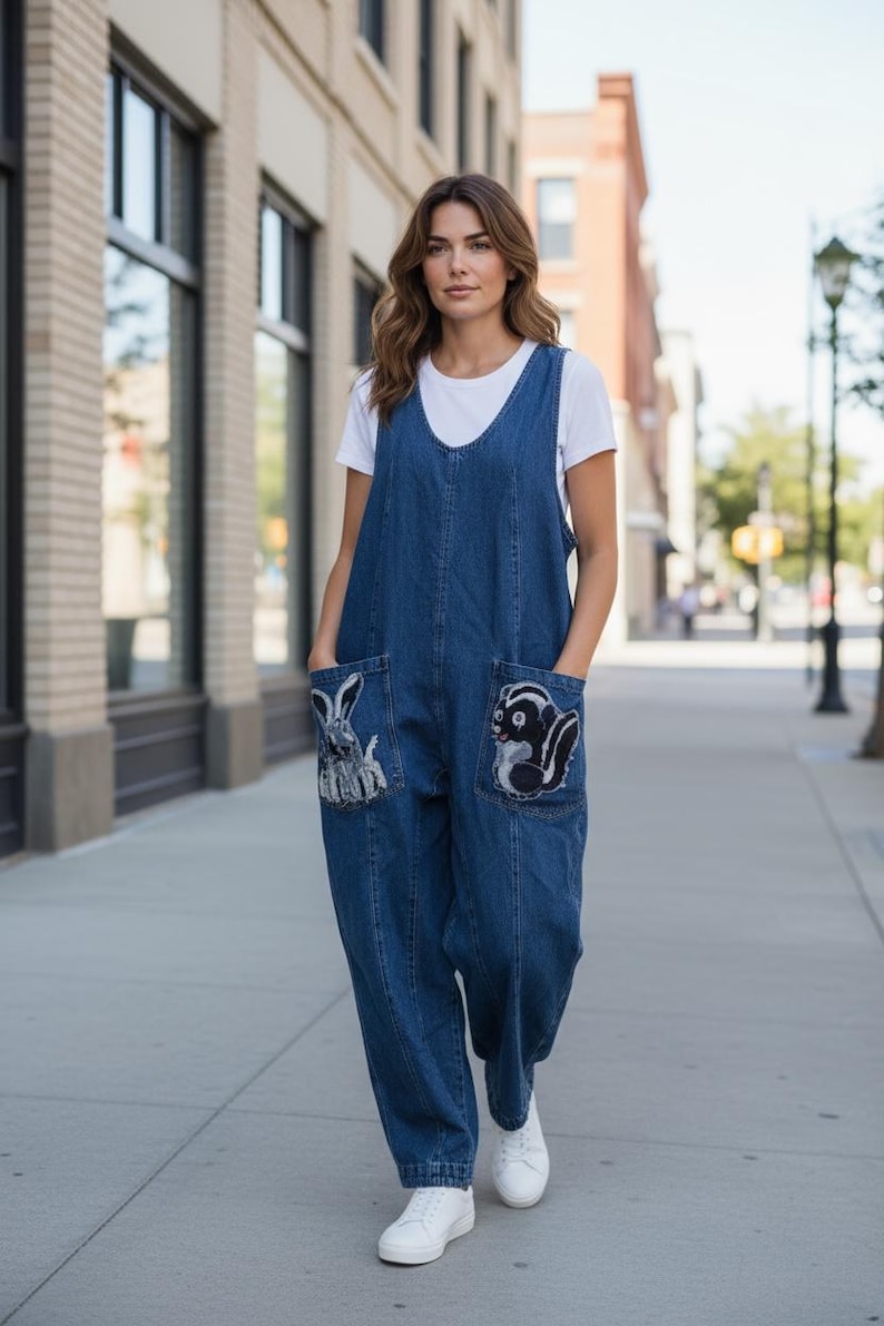 May include: A woman wearing a blue denim jumpsuit with two large front pockets. Each pocket features an embroidered animal design: a rabbit on the left and a skunk on the right. The jumpsuit is paired with a white t-shirt and white sneakers.