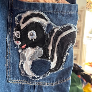 May include: Blue denim pocket with a cartoon skunk applique. The skunk is black and white with light blue eyes and a pink mouth. The applique has a frayed edge, adding a unique, handmade touch to the denim.