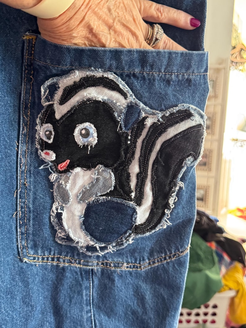 May include: Blue denim pocket with a cartoon skunk applique. The skunk is black and white with light blue eyes and a pink mouth. The applique has a frayed edge, adding a unique, handmade touch to the denim.