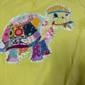 May include: A yellow sweatshirt featuring a patchwork turtle applique. The turtle is made from various coloured fabrics and has a smiling face. The sweatshirt has the text "OLD NAVY" on the front.