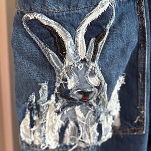 May include: Close-up of a denim pocket featuring a rabbit applique. The rabbit is constructed from various denim scraps, with a distressed, textured appearance. The rabbit has black and gray details and a small red nose.