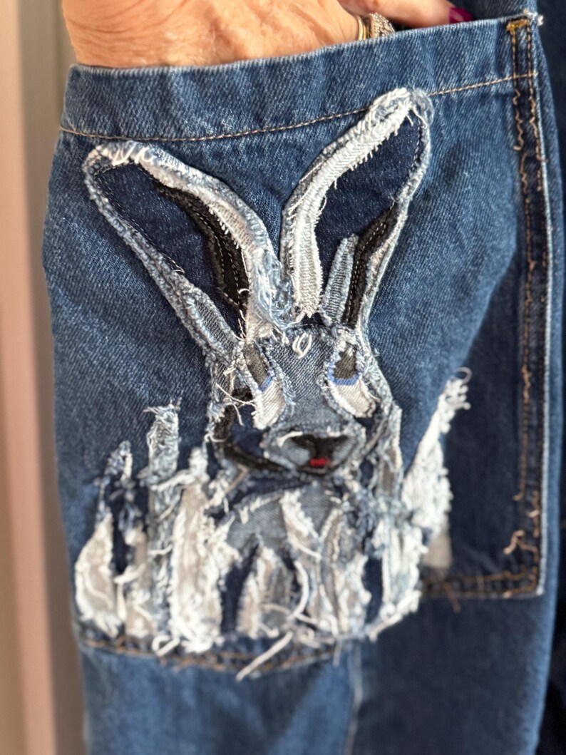 May include: Close-up of a denim pocket featuring a rabbit applique. The rabbit is constructed from various denim scraps, with a distressed, textured appearance. The rabbit has black and gray details and a small red nose.