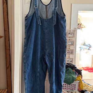May include: A pair of blue denim overalls hanging on a black hanger. The overalls have adjustable straps with metal buckles and a zipper closure on the back. The garment has pockets and visible stitching details.