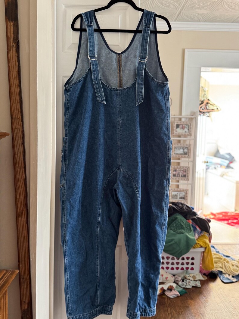 May include: A pair of blue denim overalls hanging on a black hanger. The overalls have adjustable straps with metal buckles and a zipper closure on the back. The garment has pockets and visible stitching details.
