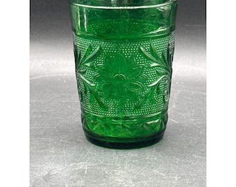 May include: A green glass tumbler with a detailed floral and dot pattern. The glass has a thick base and a straight, slightly flared rim. The design includes a central flower and surrounding leaves.