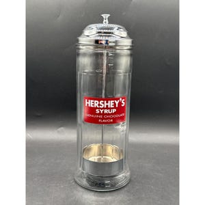 May include: A clear glass syrup dispenser with a shiny chrome lid and a matching chrome base insert. The front features a rectangular red label with white text that reads HERSHEY'S SYRUP GENUINE CHOCOLATE FLAVOR. The dispenser has a tall cylindrical shape.