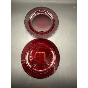 May include: Two ruby red glass plates. The plates are round with a slightly raised center and a beaded edge. The plates are a deep, rich red color and appear to be made of glass. The plates are likely for serving or display.