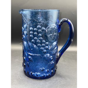 May include: A vintage cobalt blue glass pitcher with a handle and a spout. The pitcher is decorated with embossed fruit and leaf designs, including grapes and citrus slices. The pitcher is a classic piece of glassware.