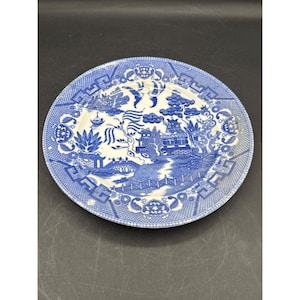 May include: A round, blue and white ceramic plate with a Willow pattern design. The plate features a detailed landscape scene with a bridge, trees, and buildings. The rim has a repeating geometric pattern. The plate is approximately 10 inches in diameter.