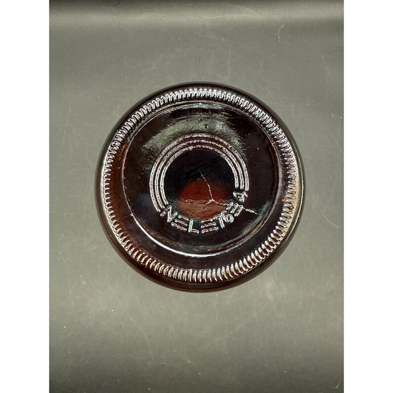 May include: A close-up shot of the bottom of a brown glass bottle. The base has a circular design with the text "NEL=76" etched into the glass. The rim of the bottle has a textured, ridged pattern.