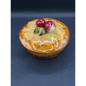 May include: A ceramic pie dish with a lid, featuring a glazed, golden-brown finish. The lid is adorned with two red apple-shaped ornaments and green leaves. The dish is round and appears to be vintage.