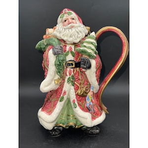May include: A ceramic Santa Claus pitcher with a red and green suit, a white beard, and a green Christmas tree in his arm. The pitcher has a gold handle and is decorated with holly and other Christmas motifs.