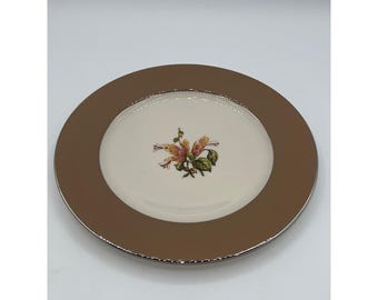 Puede incluir: Un plato de cerámica blanco con un borde marrón y un borde plateado. El plato presenta un diseño floral con flores naranjas y rosas.