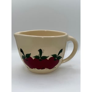 以下が含まれることがあります： クリーム色のセラミック計量カップ。取っ手と注ぎ口が付いています。カップは、緑の葉が付いた3つの大きな赤いリンゴで飾られています。リンゴはフォークアートスタイルで描かれています。