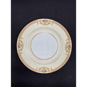 Puede incluir: Un plato de porcelana color crema con un borde floral y dorado decorativo. El plato presenta un círculo interior festoneado y diseños florales ornamentados en rosa, azul y dorado. El plato está sobre un fondo negro.