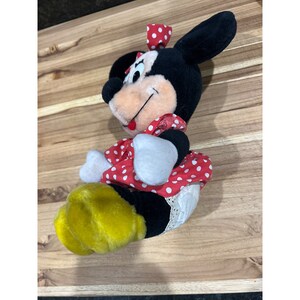 Pu&ograve; includere: Un giocattolo di peluche di Minnie Mouse che indossa un vestito rosso con pois bianchi e un fiocco bianco con pois rossi. Il giocattolo ha scarpe gialle e un bordo di pizzo bianco sull'abito.