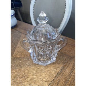 Puede incluir: Un azucarero de vidrio transparente con tapa. La tapa tiene un pomo en forma de diamante en la parte superior. El tazón tiene un borde festoneado y un patrón decorativo en los lados.