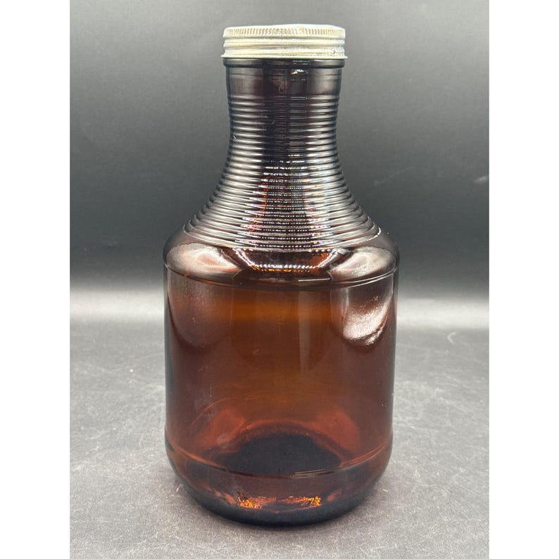 May include: A vintage brown glass bottle with a ribbed neck and a silver metal lid. The bottle has a rounded base and a cylindrical body. The glass has a slight amber tint, and the bottle appears to be empty.