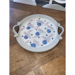 Puede incluir: Una bandeja de servir de cerámica blanca con dos asas. La bandeja tiene un patrón geométrico rosa y azul con pequeños puntos azules.