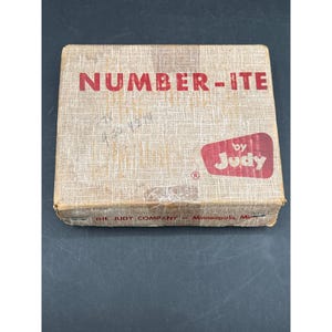 Puede incluir: Una caja vintage para "NUMBER-ITE" de Judy, con una superficie beige texturizada y letras rojas. La caja tiene forma rectangular con un logotipo rojo y el texto "THE JUDY COMPANY - Minneapolis, Minn." impreso en la parte inferior.