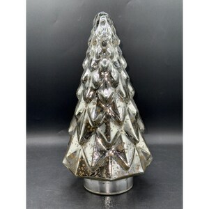 May include: A silver-colored, decorative Christmas tree with a textured, faceted design. The tree has a metallic finish and sits on a round, silver base. The tree's shape is a classic Christmas tree silhouette.