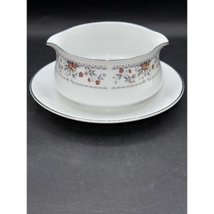 May include: White porcelain gravy boat and saucer set. The gravy boat features a floral pattern with orange, blue, and brown details, and a silver trim. The saucer also has a silver trim. Ideal for serving gravy or sauces.