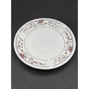 May include: White ceramic saucer with a floral pattern in red, blue, and grey. The saucer has a central indentation and a decorative border with small flowers and leaves. A delicate, elegant design.
