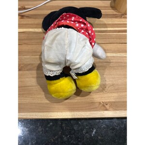 Pu&ograve; includere: Un giocattolo di peluche di un personaggio dei cartoni animati che indossa un vestito rosso e bianco a pois e pantaloni bianchi con bordini neri. Il giocattolo ha scarpe gialle e occhi neri.