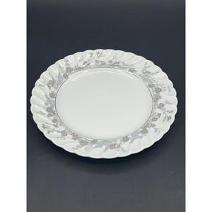 May include: A white ceramic dinner plate with a silver floral design around the rim. The plate has a slightly scalloped edge.