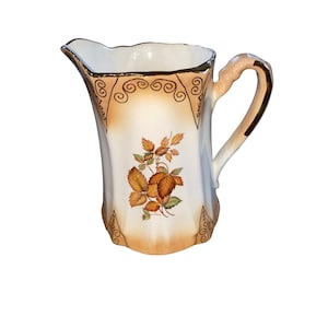Pode incluir: Um jarro de cerâmica branca com uma borda dourada e um desenho floral com folhas marrons. O jarro tem uma alça e um bico arredondado.