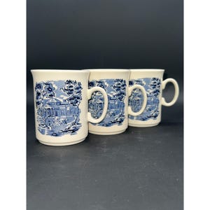 Peut inclure: Trois tasses en céramique blanche avec des motifs bleus et blancs. Chaque tasse présente une illustration bleue et blanche d'une maison avec des arbres et une calèche. Les tasses ont une forme arrondie et une poignée.