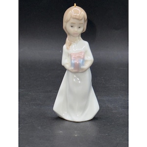 May include: A porcelain figurine of a young girl in a white dress, holding a small gift box with a pink bow. The girl has blonde hair and a serene expression. A gold loop is at the top of the figurine.