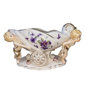 May include: A white ceramic centerpiece with a shell-shaped bowl, adorned with purple floral designs and gold accents. Two cherubic figures support the bowl, which rests on a decorative wheel base. A charming decorative piece.