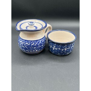 May include: A ceramic creamer and sugar bowl set. The creamer has a handle, spout, and lid. Both pieces are off-white with a blue floral pattern and blue trim. The sugar bowl is round and open.