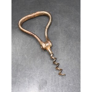May include: A vintage metal corkscrew featuring a triangular-shaped handle with a weathered, brownish-grey patina. The tool consists of a hinged loop handle connected to a central pivot point, extending into a spiraled metal worm for extracting corks.