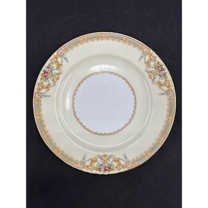 Puede incluir: Plato vintage color crema con borde festoneado y círculo blanco central. El plato presenta un borde dorado y verde azulado con diseños florales y de volutas. El plato decorativo es probablemente para servir o exhibir.