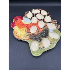 Puede incluir: Un soporte para huevos de cerámica con forma de gallo, con cresta roja y plumas naranjas y negras. El soporte tiene espacios para una docena de huevos, con una base verde y una sección marrón con forma de madera.
