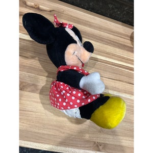 Pu&ograve; includere: Un giocattolo di peluche nero e bianco di Minnie Mouse che indossa un vestito rosso con pois bianchi e un fiocco giallo. Il giocattolo &egrave; seduto su una superficie di legno.