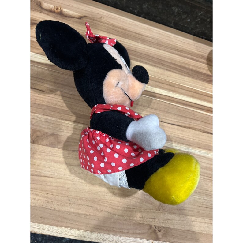 Pu&ograve; includere: Un giocattolo di peluche nero e bianco di Minnie Mouse che indossa un vestito rosso con pois bianchi e un fiocco giallo. Il giocattolo &egrave; seduto su una superficie di legno.
