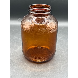 Op de afbeelding: Amberkleurige glazen pot met een brede opening en een getextureerd oppervlak. De pot heeft een cilindrische vorm met een afgeronde basis en een getextureerd, gestippeld patroon op het bovenste gedeelte. De pot is leeg en staat op een grijs oppervlak.