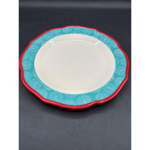 May include: A white ceramic plate with a scalloped edge. The rim is red, with a turquoise band featuring a white floral pattern. The plate is designed for serving food.