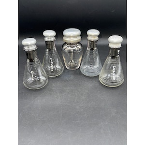 May include: A set of five vintage glass condiment bottles with silver-colored tops and black accents. The bottles are labeled "Oil", "Vinegar", "Salt", and "Pepper". The central bottle is a shaker. The bottles are arranged on a dark surface.