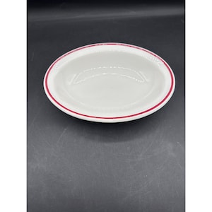 May include: White oval serving bowl with a red rim. The bowl is empty and appears to be made of ceramic or porcelain. The bowl is centered in the frame against a dark background.