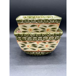 Może przedstawiać: Dwie ceramiczne miski ułożone jedna na drugiej. Miski są kwadratowe z zaokrąglonymi rogami i kremowym tłem. Ozdobione są zielonymi, brązowymi i żółtymi wzorami kwiatowymi. Obręcze są zielone.