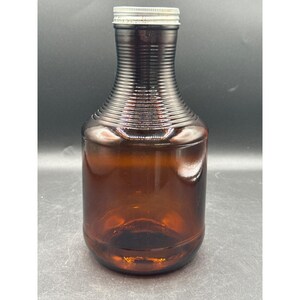 May include: A brown glass bottle with a ribbed neck and a silver metal cap. The bottle has a rounded body and a dark amber color, suggesting it may be used for storing liquids.