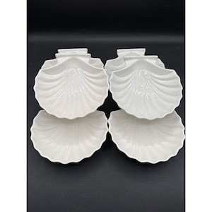 以下が含まれることがあります： 白い貝殻型の陶器の皿が6つ。各皿はスカラップの縁と、自然の貝殻に似た、詳細なリブ付きの表面を持っています。皿は、暗い背景に対して、3つずつ2列に配置されています。